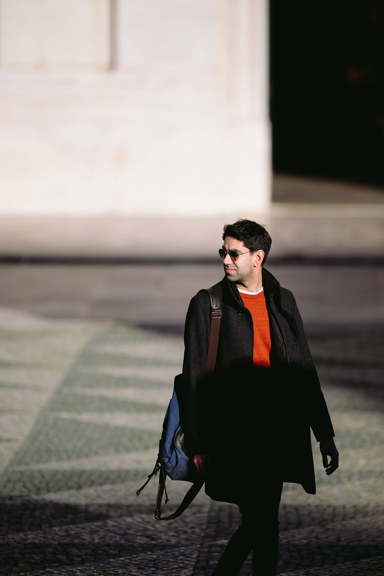 Man in sunglasses, black coat and orange pullover crossing the square in front of the Lisbon City Hall just before the sunset