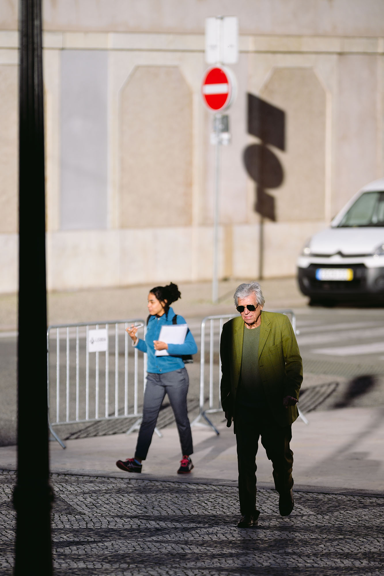 Woman in blue jacket, man in green blazer and sunglasses crossing the square in front of the Lisbon City Hall just before the sunset
