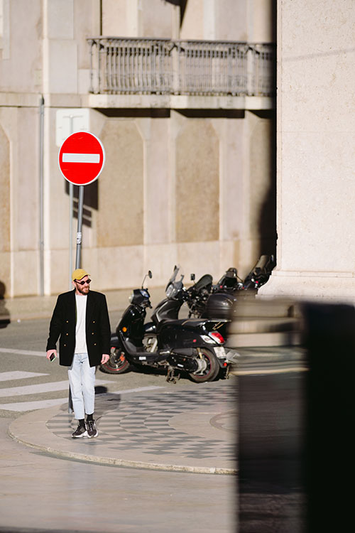 Man in yellow cap, sunglasses, white t-shirt and black blazer about to cross the square in front of the Lisbon City Hall just before the sunset