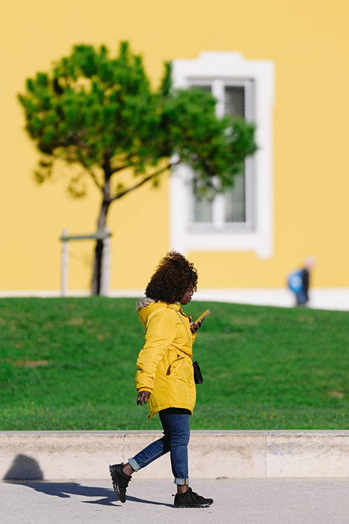 Woman in the yellow jacket walking on a sunny day in front of the yellow wall and green tree in Lisbon, Portugal